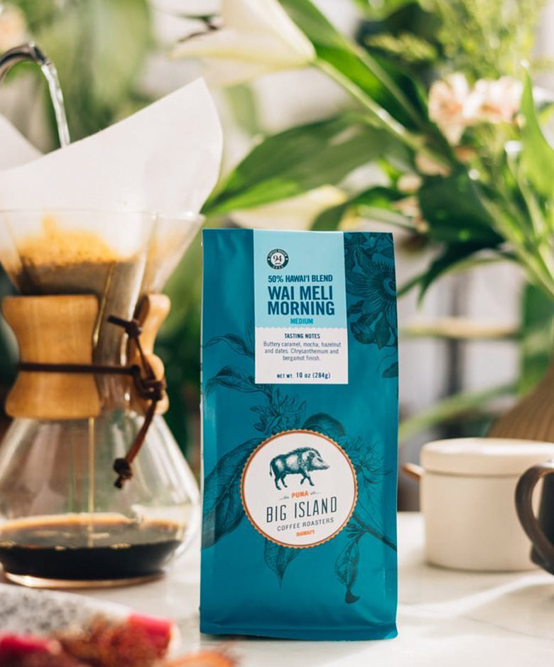 A bag of Big Island Coffee Roasters Wai Meli Morning coffee stands on a table in a bright, real environment with a glass pour-over coffee maker, flowers, and a mug blurred in the background.