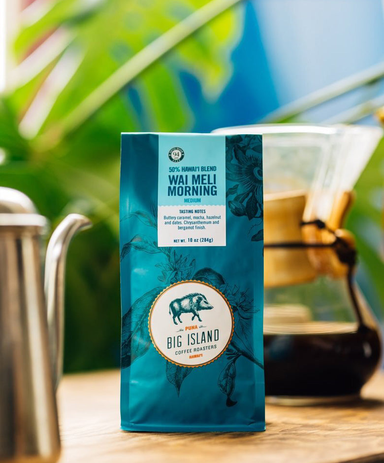 A bag of Big Island Coffee Roasters Wai Meli Morning coffee stands on a wooden table with a coffee pour-over setup and blurred tropical greenery in the background.